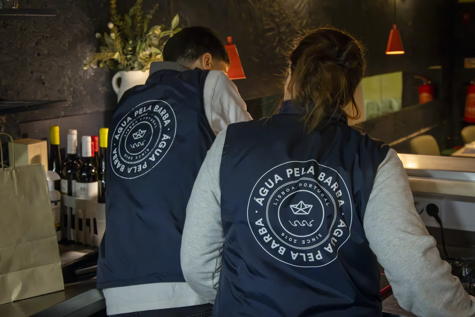 Dois empregados de costas, ao balcão, com a marca do restaurante nos coletes